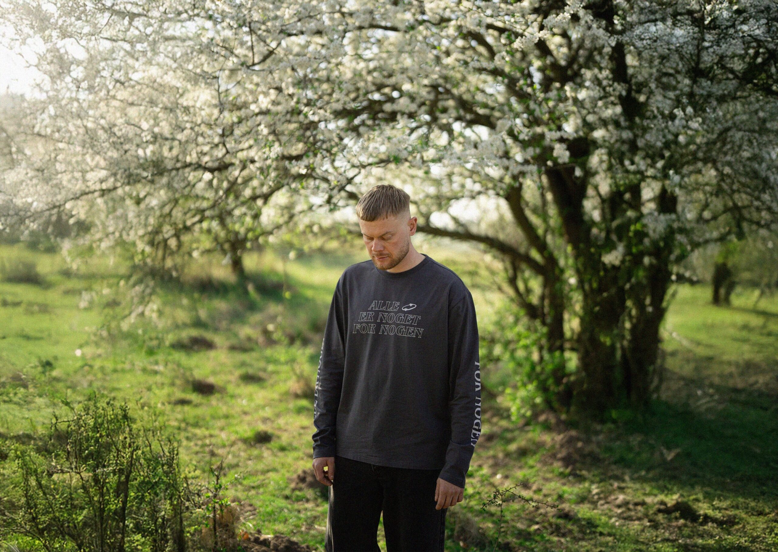 Billede af artisten URO, der står i en skov foran et stort træ med hvide blomster og kigger ned mod græsset.